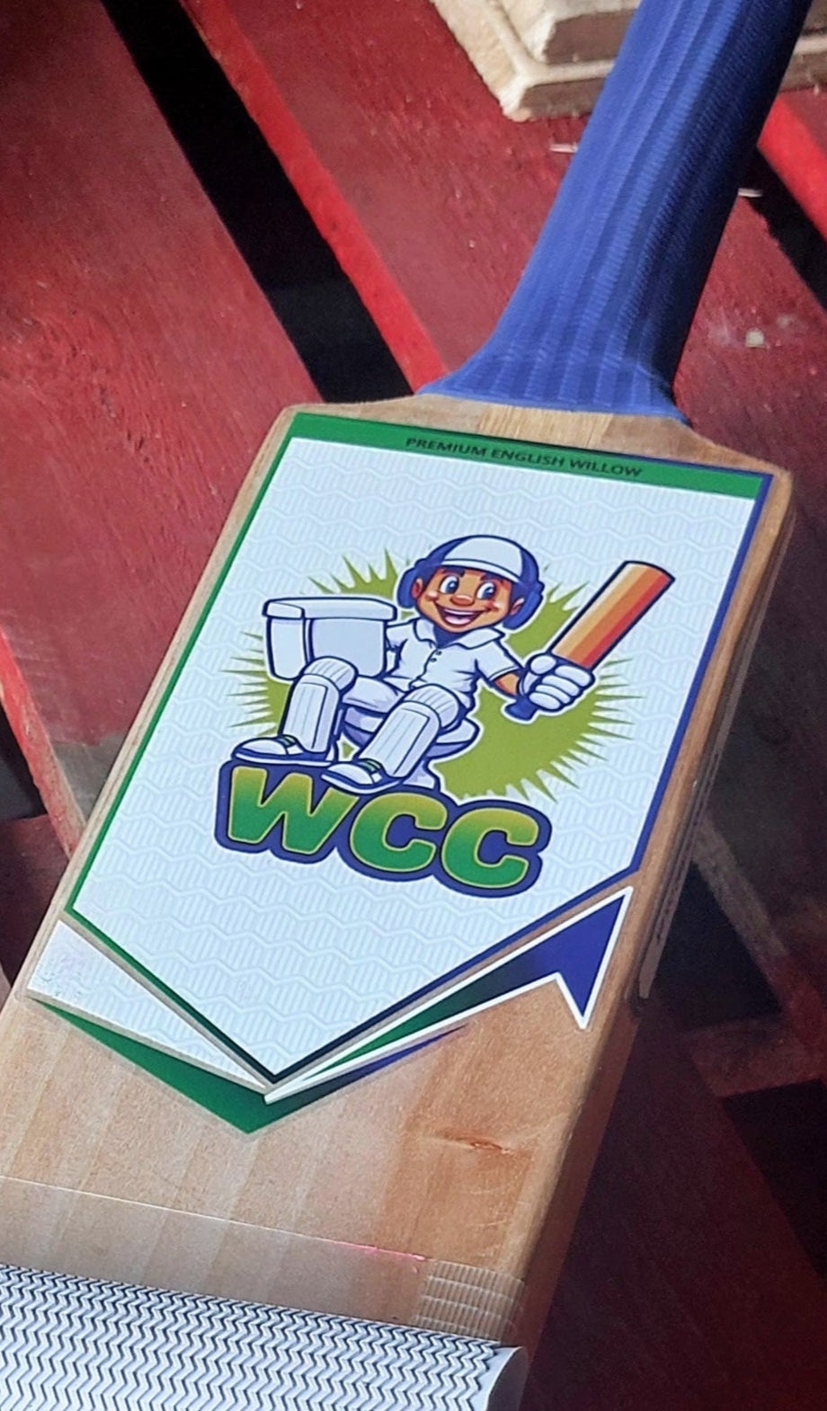 WC Cricket cricket bat restoration showing WCC stickered blade and blue grip on wooden bench for cricket bat sticker removal